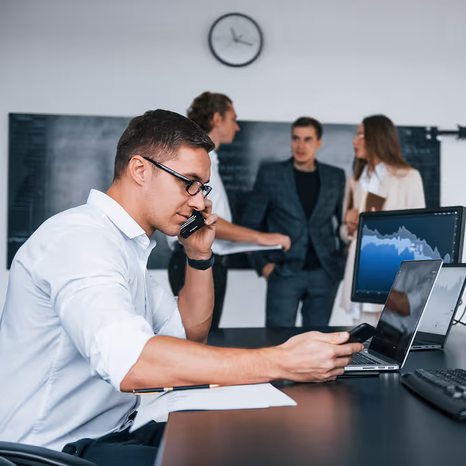 Man in glasses talking on the phone while working on a laptop in an office with coworkers and a clock in the background.