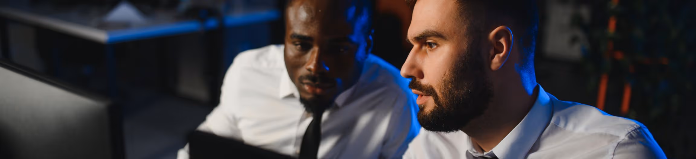 Two men in white shirts and ties focused on a laptop screen in a dimly lit office.