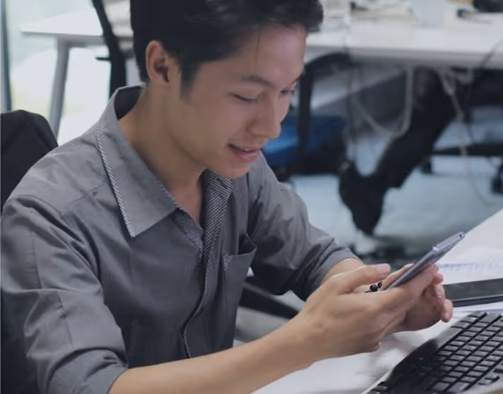 Young man smiling and looking at his smartphone while sitting at a desk with a keyboard and papers.